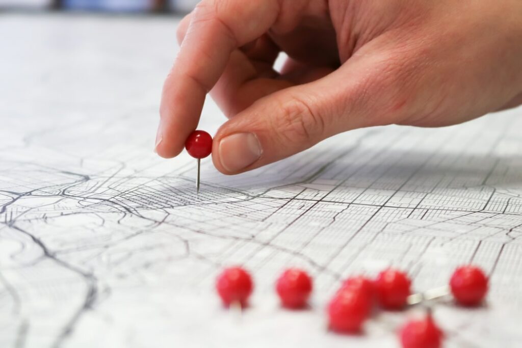 A white hand places a red pin on a white map with black lines. A collection of more red pins lay off to the right side.